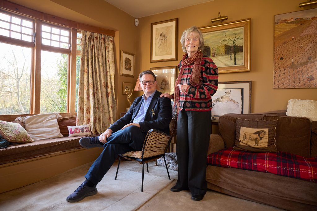 Will Travers is sat in a chair, with his mother Dame Virginia McKenna stood at his side, with a hand on his shoulder. Both are smiling at the camera.