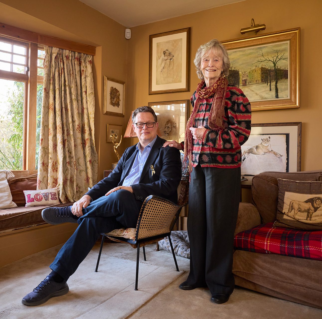 Will Travers is sat in a chair, with his mother Dame Virginia McKenna stood at his side, with a hand on his shoulder. Both are smiling at the camera.