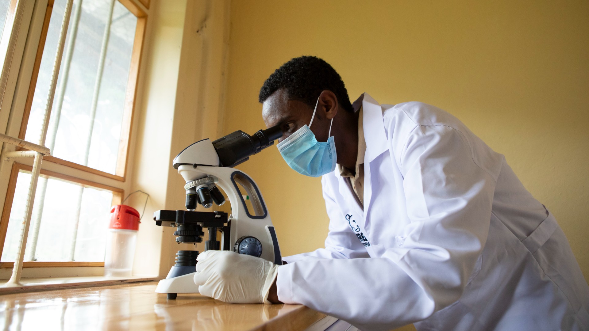 A man wearing a white lab coat and face mask, looking at samples through a microscope