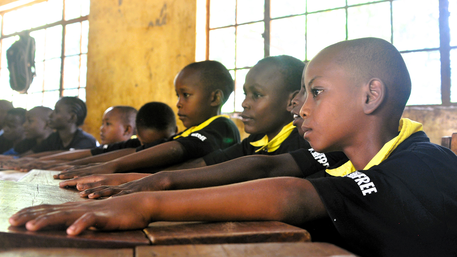 A line of Kenyan school children sit at desks, with one arm reaching forward across their desks