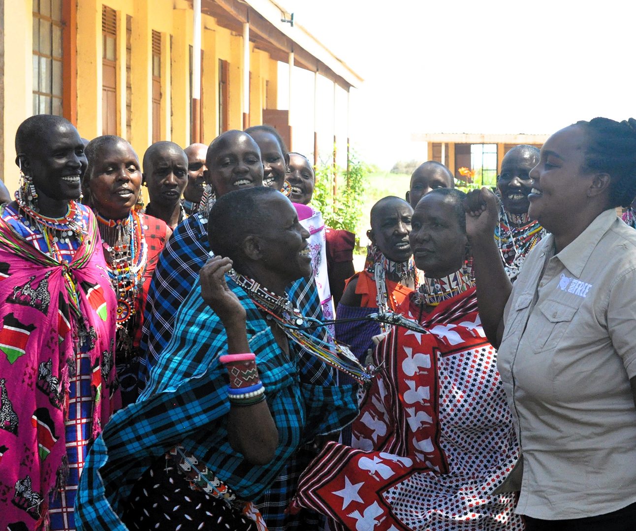 A Kenyan woman in traditional dress is leaning forward and dancing with a woman in Born Free uniform, in front of a crowd