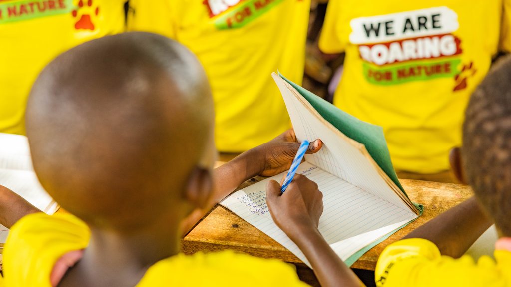 A child facing away from the camera is writing in a text book, surrounded by other students wearing Born Free conservation club Tshirts