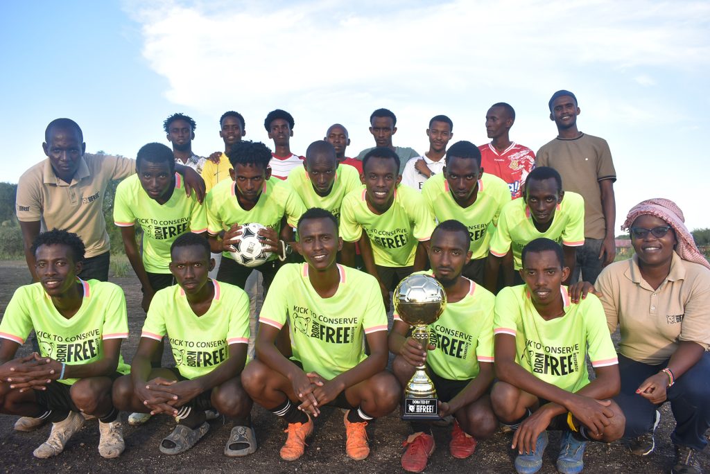 A team of male Kenyan football players pose for a photo holding a football and a trophy, all are wearing Born Free T-shirts