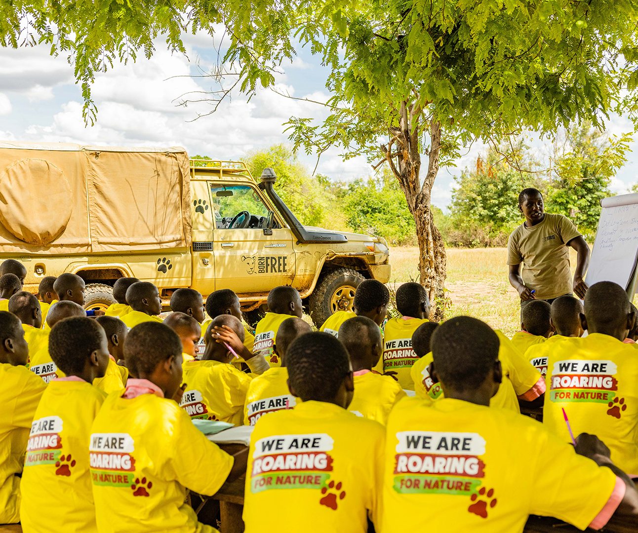 A large group of Kenyan school cildren sit with their backs to camera, facing a man in a Born Free shirt stood next to a flip chart. The children are all wearing matching yellow T-shirts with 'We are roaring for nature' logos on the back