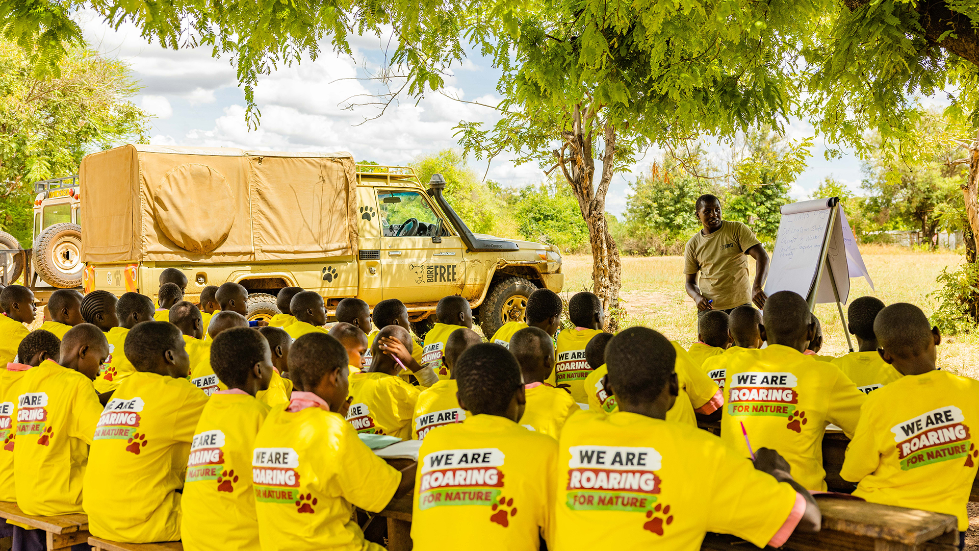 A large group of Kenyan school cildren sit with their backs to camera, facing a man in a Born Free shirt stood next to a flip chart. The children are all wearing matching yellow T-shirts with 'We are roaring for nature' logos on the back