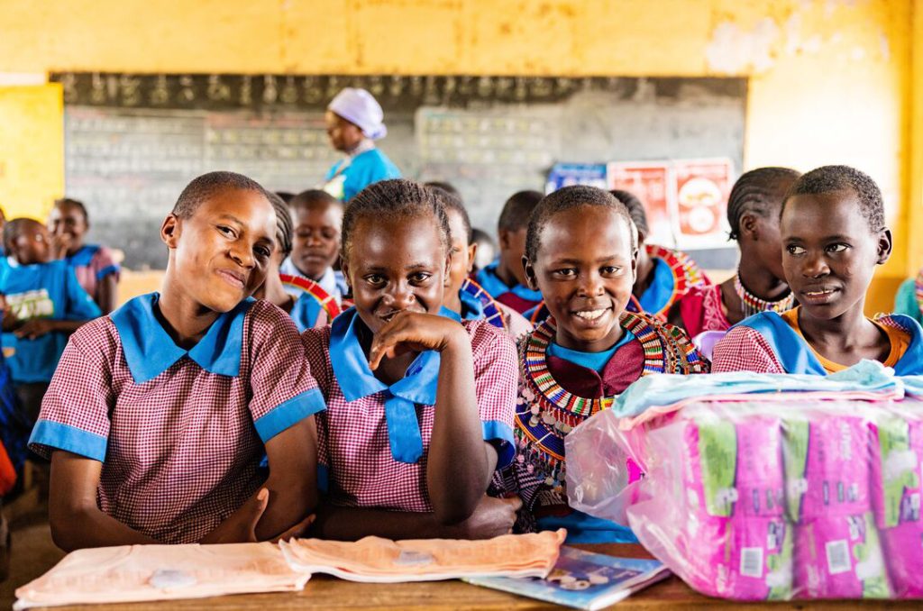 A group of four Kenyan schoolgirls smile at the camera, with a multipack of sanitary pads on the table in front of them