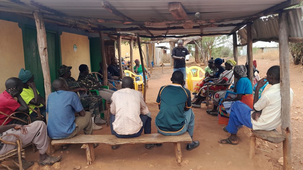 A group of Kenyan community members sit on benches, with a Born Free staff member in black T-shirt stood in front of them presenting