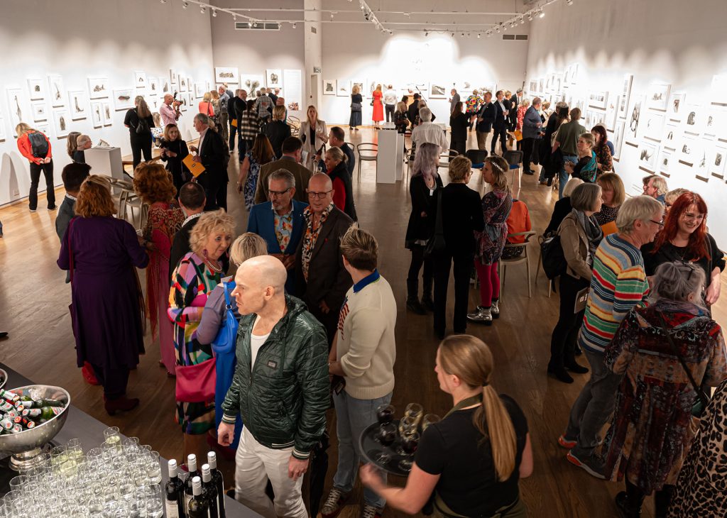 Photo taken from above of a room filled with people and pencil drawings framed on the walls