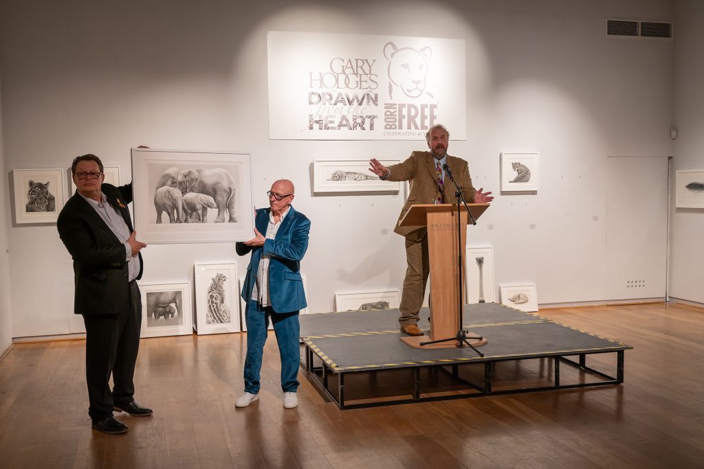 Auctioneer James Lewis stands in front of a wall of framed animal drawings; Will Travers OBE and Gary Hodges hold up one drawing