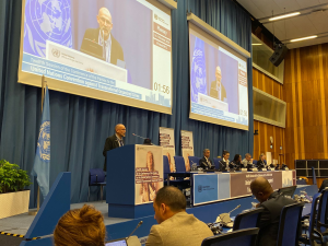 A photo showing speakers at a UN conference, with screens behind them. Mark Jones is making a speech at the podium.