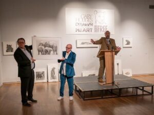 Two men holding up a drawing in an art gallery while a third man conducts an auction.