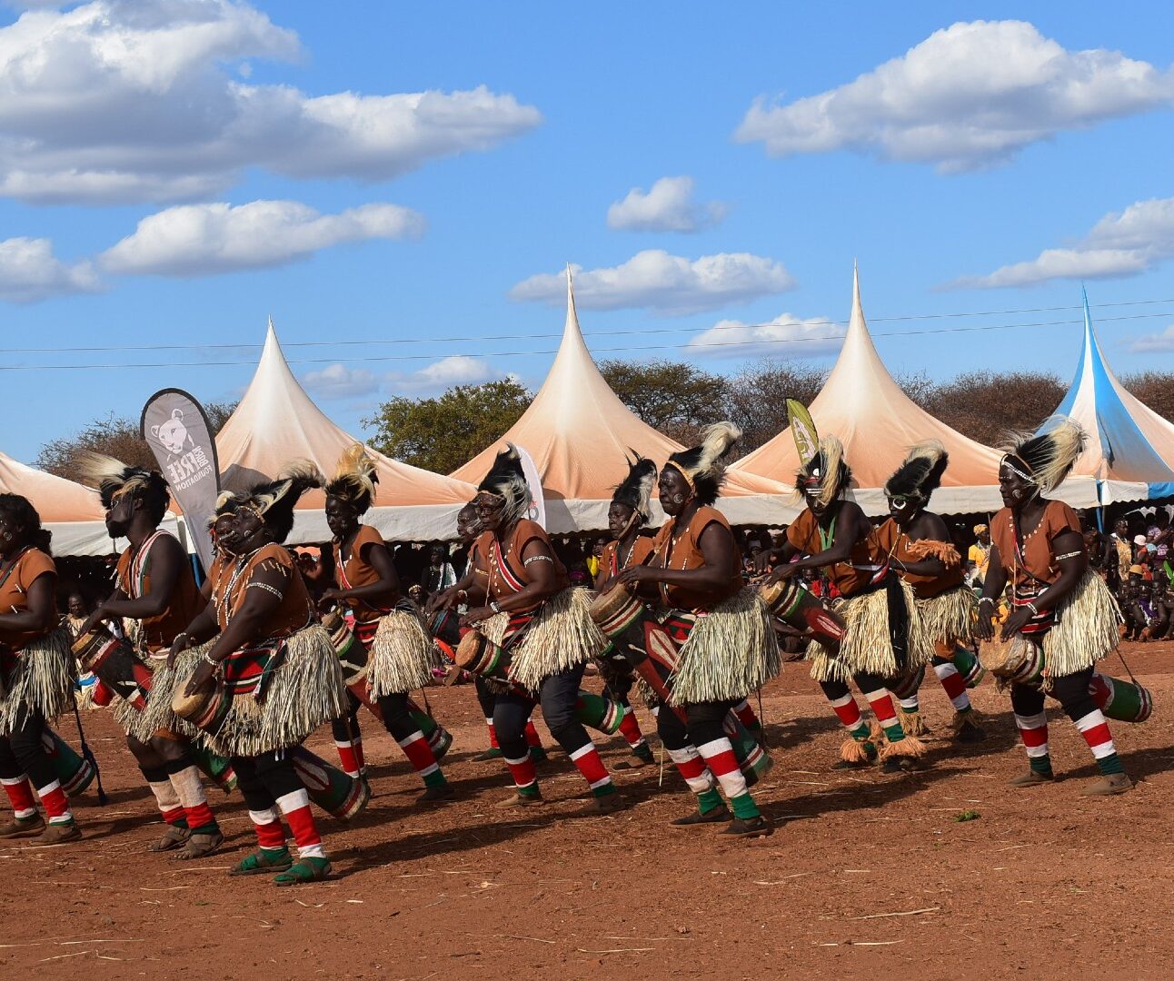 A group of men wearing traditional Kenyan dress, dancing outdoors with a blue sky in the background